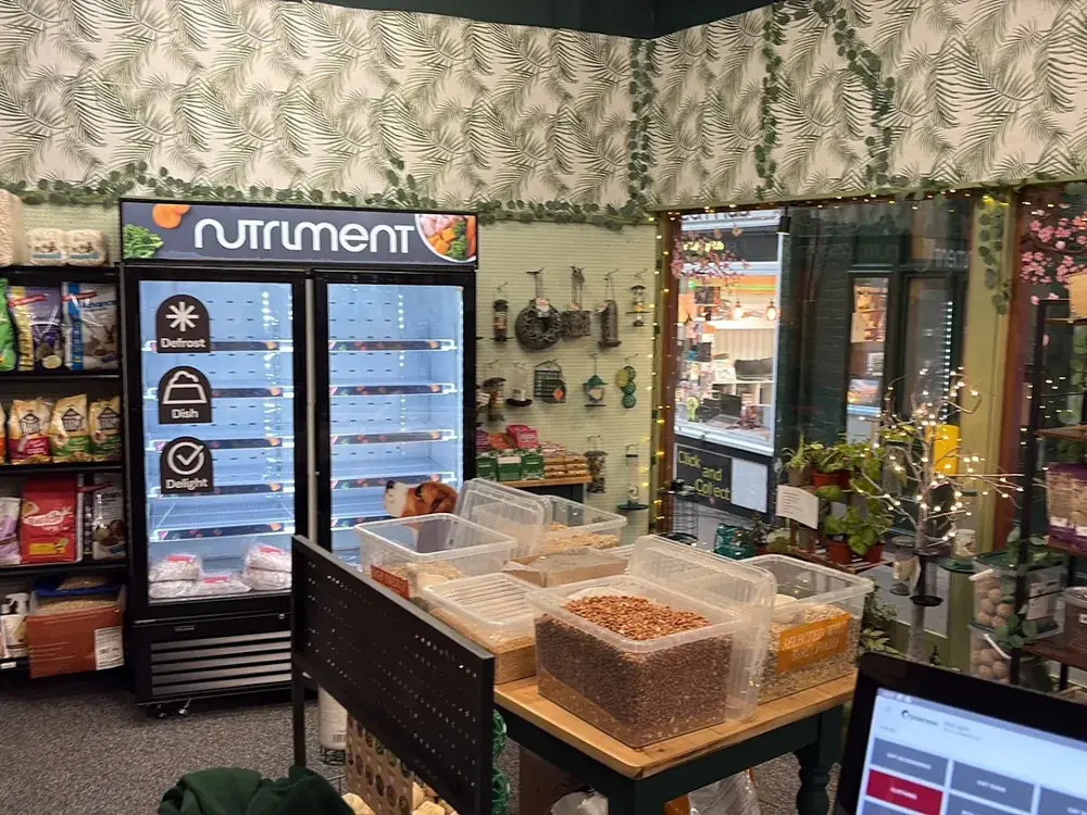 Wider view of Paws'n'Moors — Nutriment freezer left, loose-bin grain and seed mixes on a wooden table, leaf-trim jungle wallpaper.